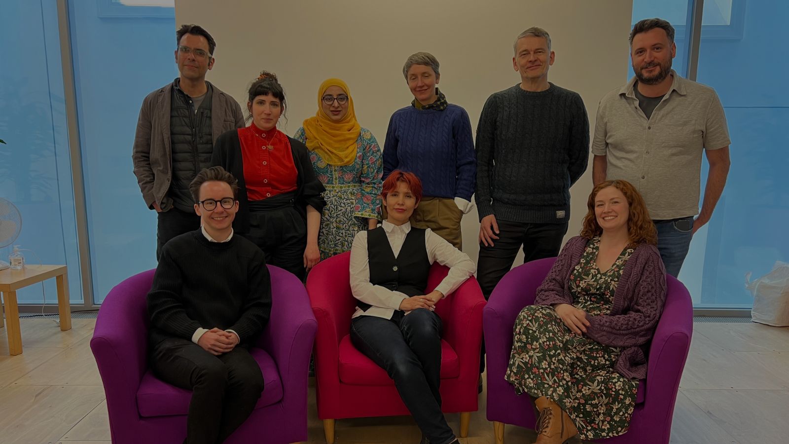 A group of people stand together. There are six people standing up, in front of them are three people sitting on chairs. They are a range of ages.