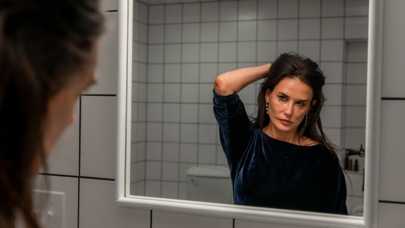 Still from the Substance, which shows actress Demi Moore looking into a mirror, lifting her hair up. She wears a navy velvet dress.