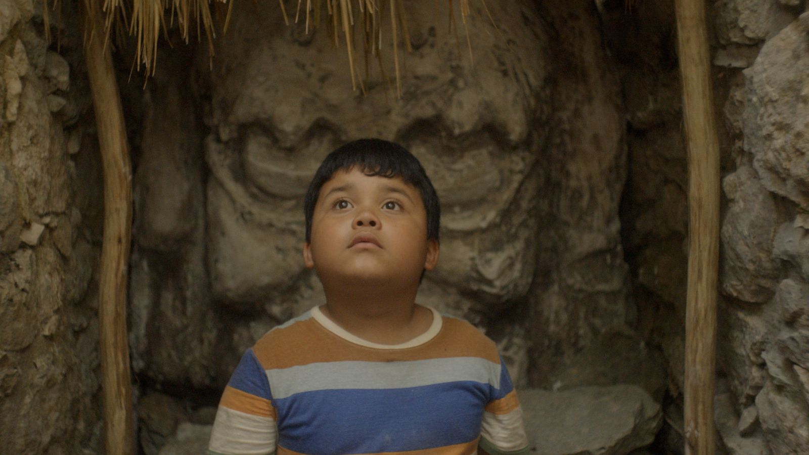 Still from Xibalba Monster, which shows a boy who wears a striped t-shirt sitting in front of a carved face in the stone.