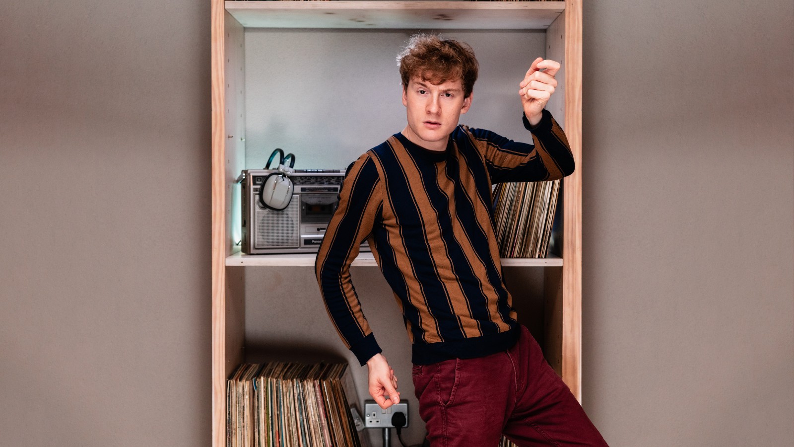 James Acaster stands in front of shelves containing records and a radio. He wears red trousers and an orange and black striped top.
