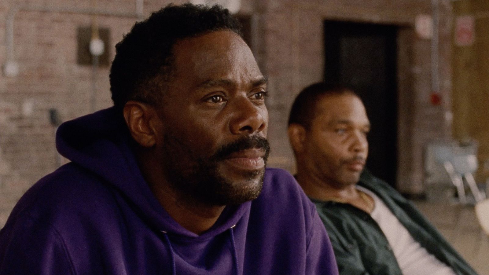 Still from Sing Sing, showing a close up of Colman Domingo, who wears a purple hoodie. Beside him another man sits wearing a white t-shirt and green overshirt. They are in a room with brick walls. Courtesy of EIFF
