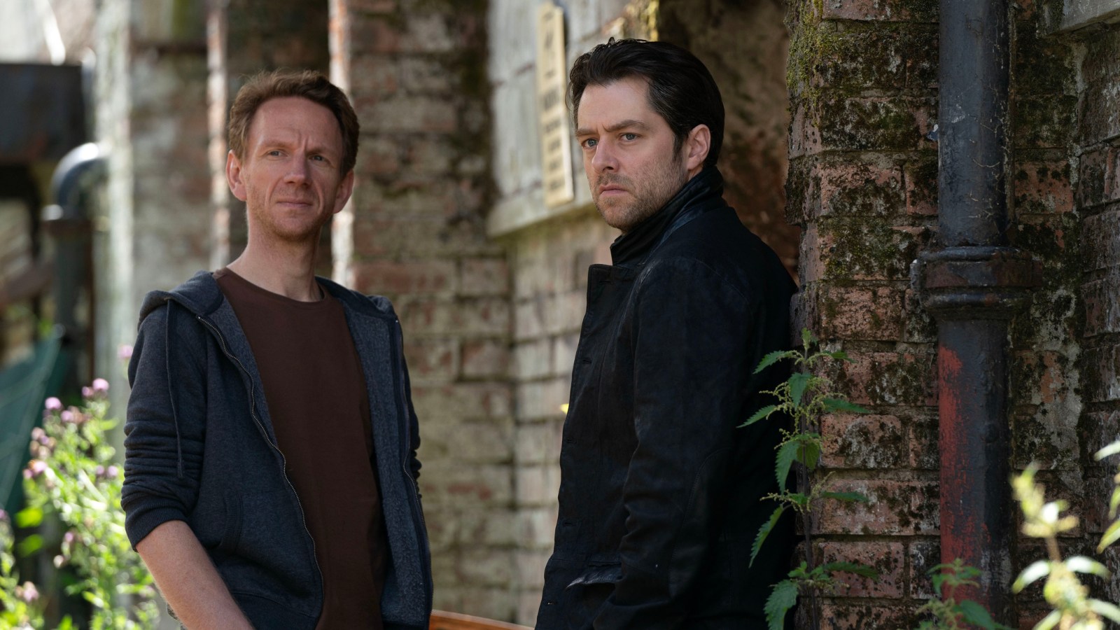 Still from Rebus, courtesy of BBC. Brian Ferguson and Richard Rankin as Michael and John Rebus, both stand in front of a brick house.