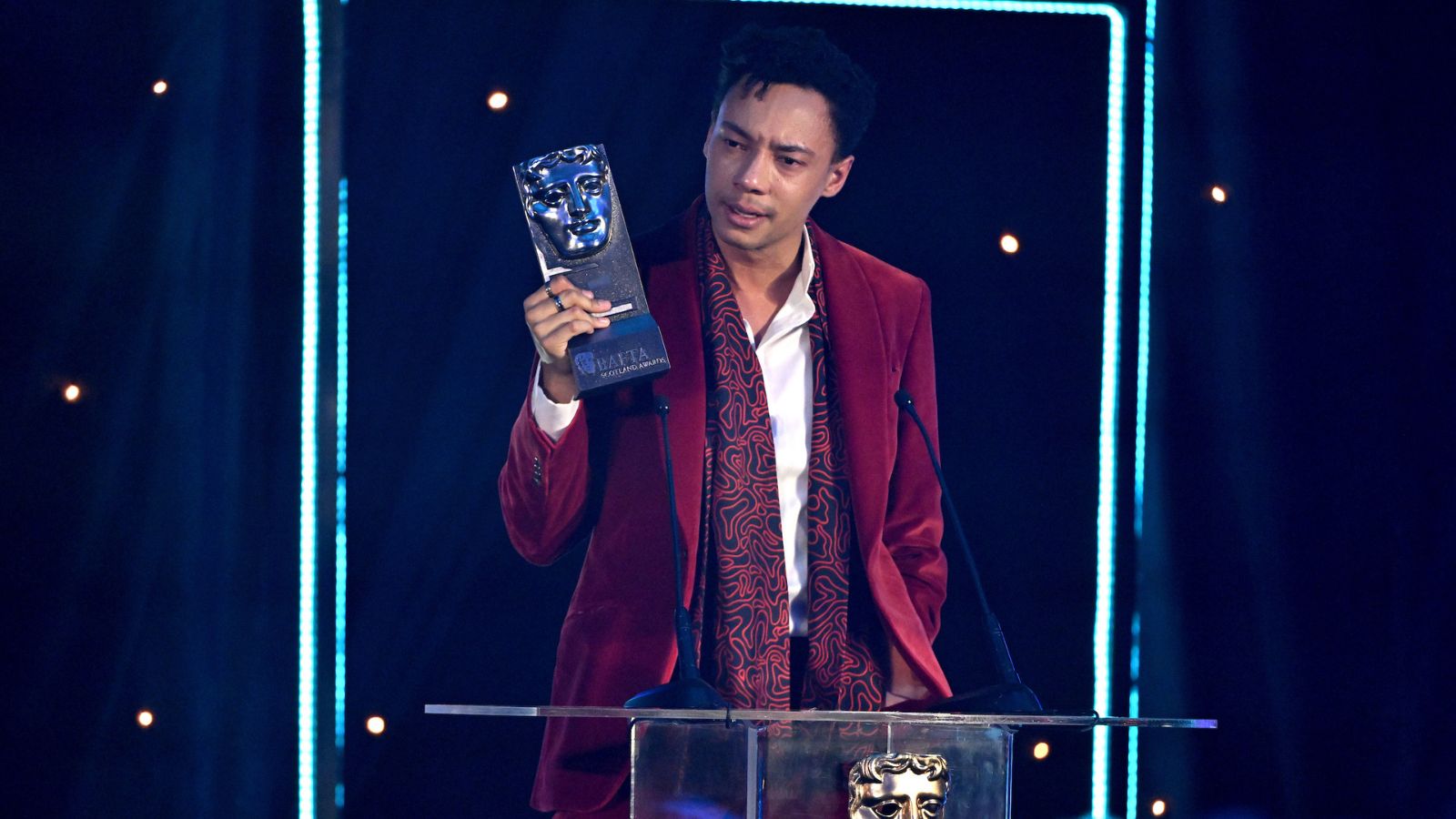 Actor Kit Young stands on stage at the BAFTA Scotland Awards with his BAFTA award. He wars a red velvet suit jacket and red and black scarf.