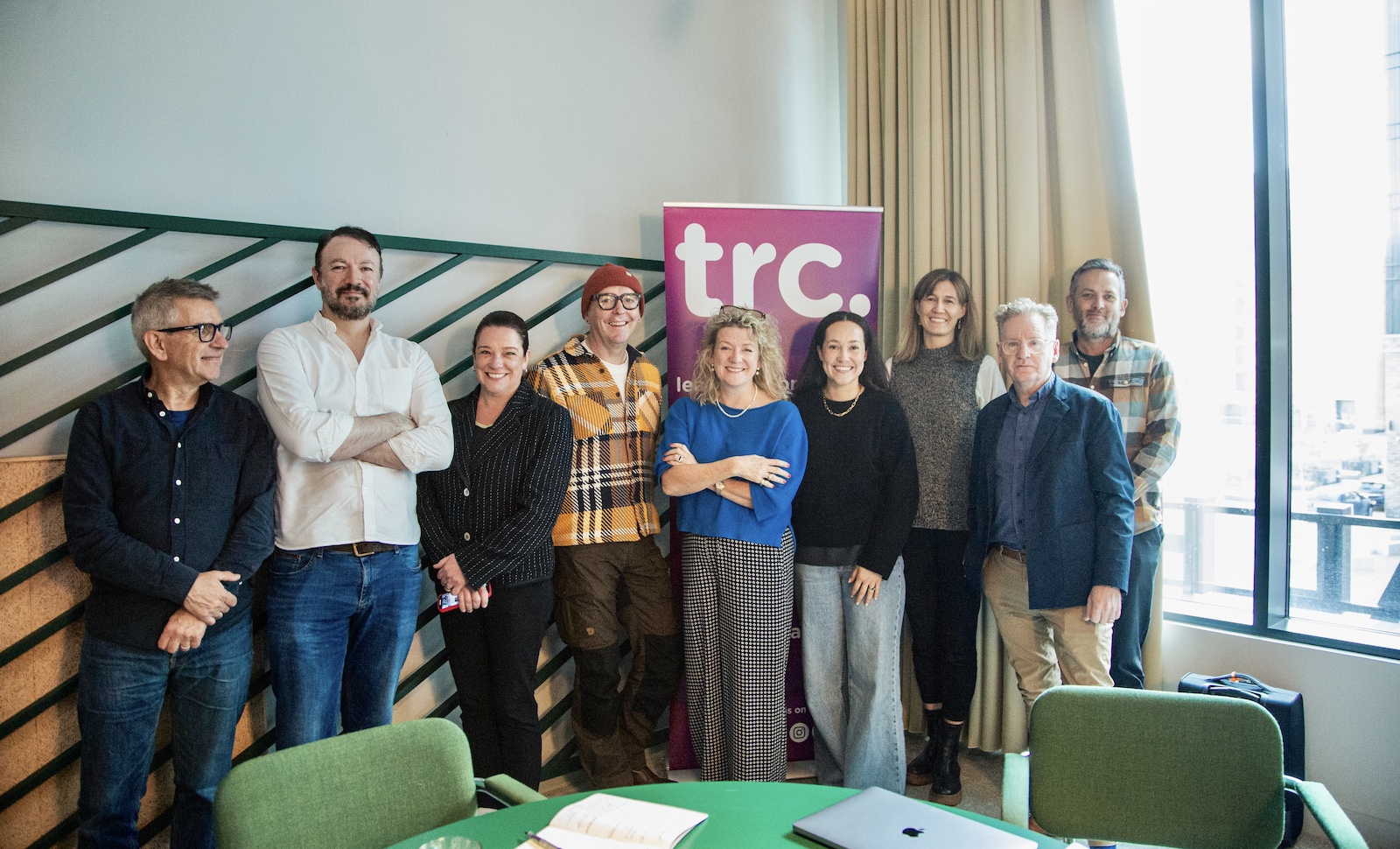 A group of people stand in a room looking cheerfully at the camera in front of a sign that says TRC - these are the representatives of the latest international horizons scripted cohort