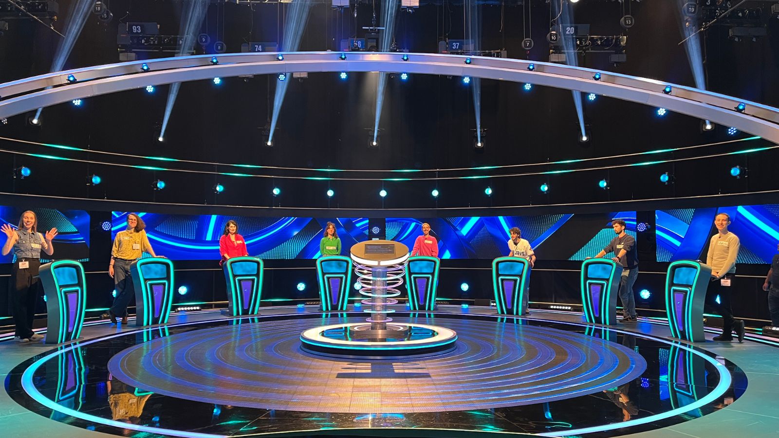 Eight people stand behind individual podiums on the set of The Weakest Link in a TV studio.