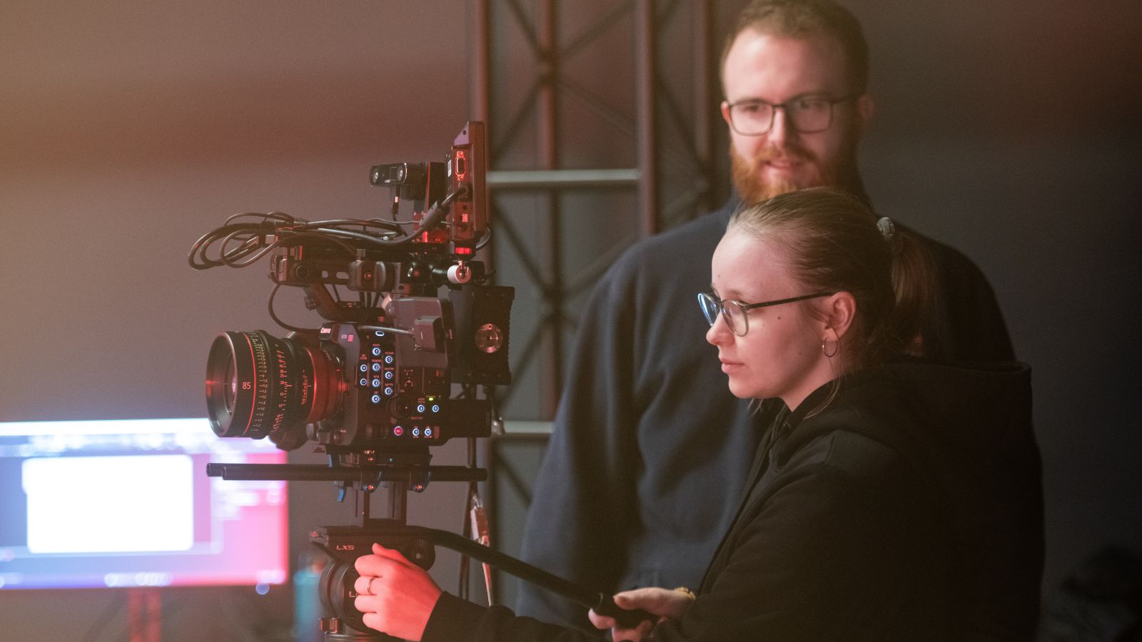 A young woman operates a camera in a studio while a man with a beard supervises