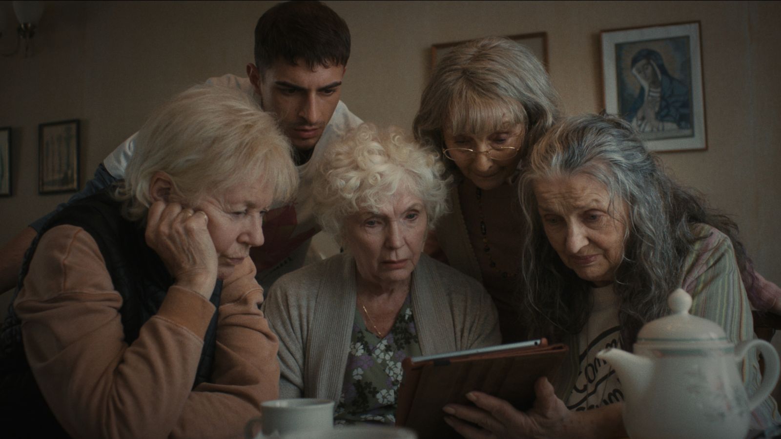 Still from Four Mothers, which shows four older women and a younger man gathered at a table together, looking at an iPad