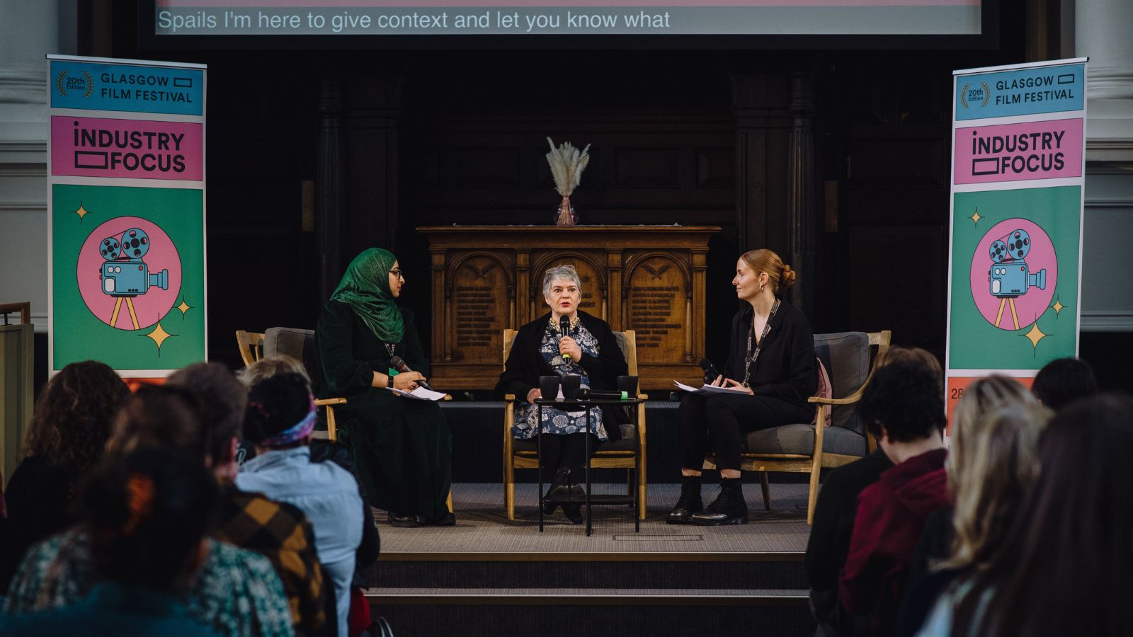 An industry event at the 2024 Glasgow Film Festival shows three women sitting on a stage having a discussion. A roller banners with the film festival logo is on either side of them.