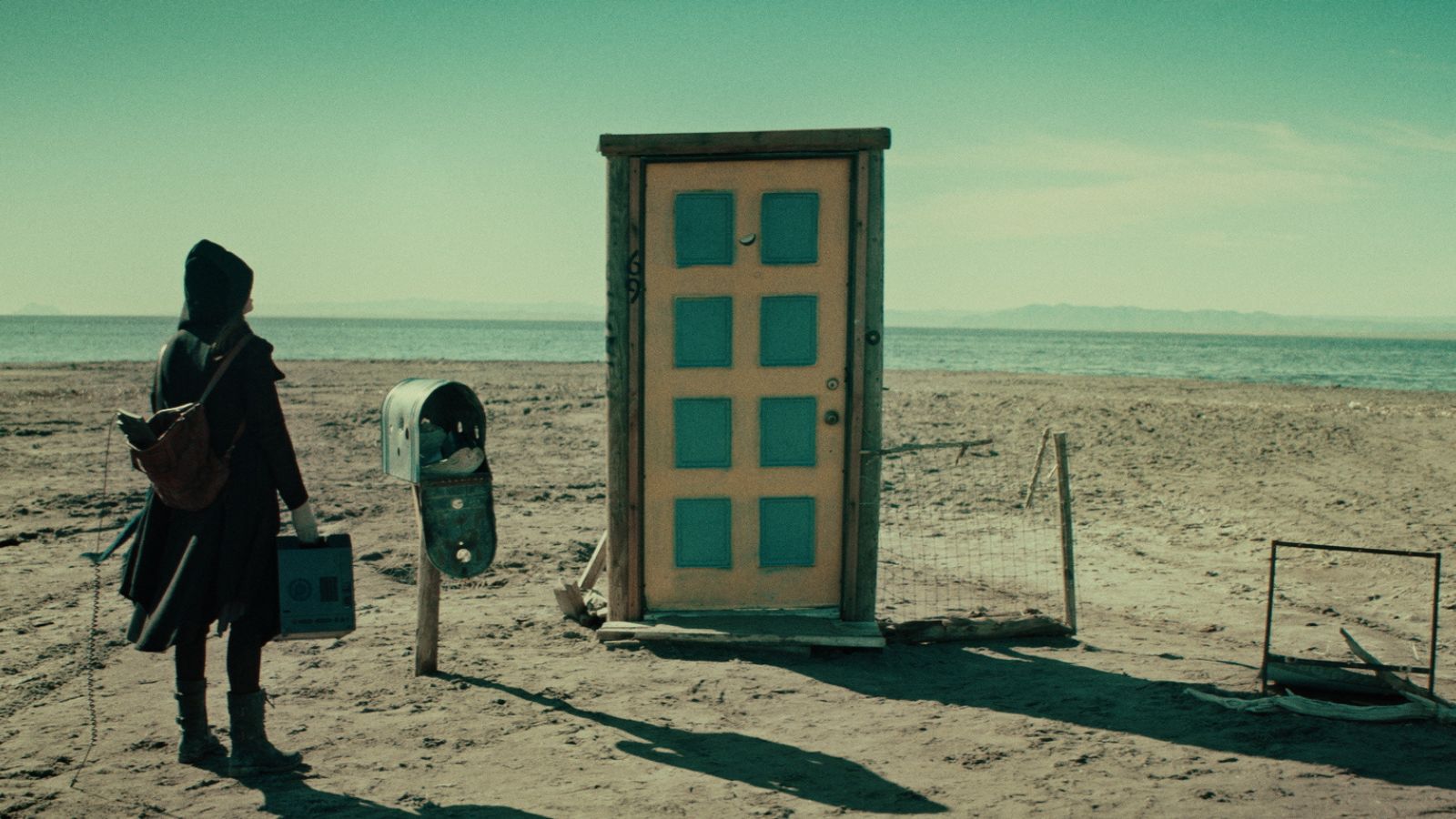 Still from Psyche, which shows a figure wearing a cloak standing in a desert landscape in front of a door, which is not attached to anything.