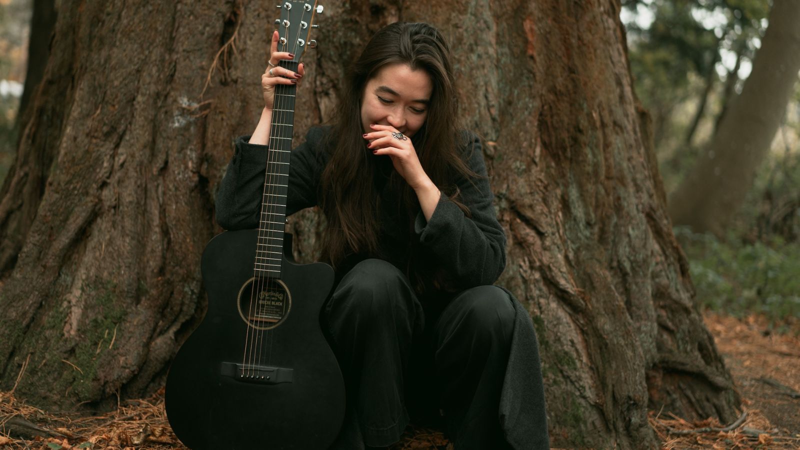 A woman sits outside against a large tree. She holds an acoustic guitar beside her.